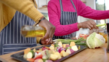 Mid-section-of-african-american-couple-in-aprons-pouring-oil-on-vegetables-in-kitchen,-slow-motion