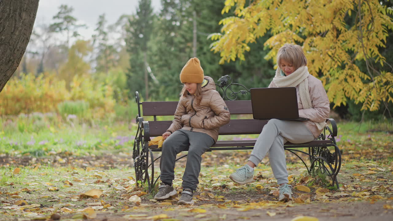 woman sits bench in autumn park working laptop while little girl plays foliage on ground beside her creating warm mother child outdoor moment with gentle interaction and subtle focus shift