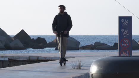 Young-male-riding-electric-scooter-on-seafront-promenade-early-morning