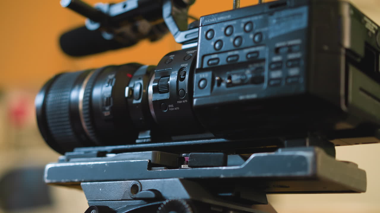 Close-up of hand adjusting camera lens settings on tripod. Focus on camera controls and lens. Background features a soft, blurred orange wall. Cinematic studio setup with detailed focus adjustment