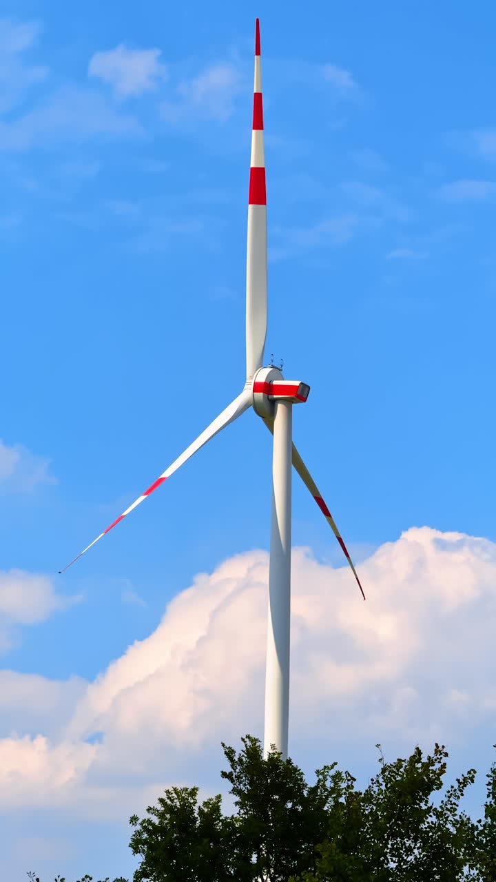 Tall wind turbine, blue sky. A large wind turbine with red and white accents spins in the breeze under a clear blue sky with fluffy clouds