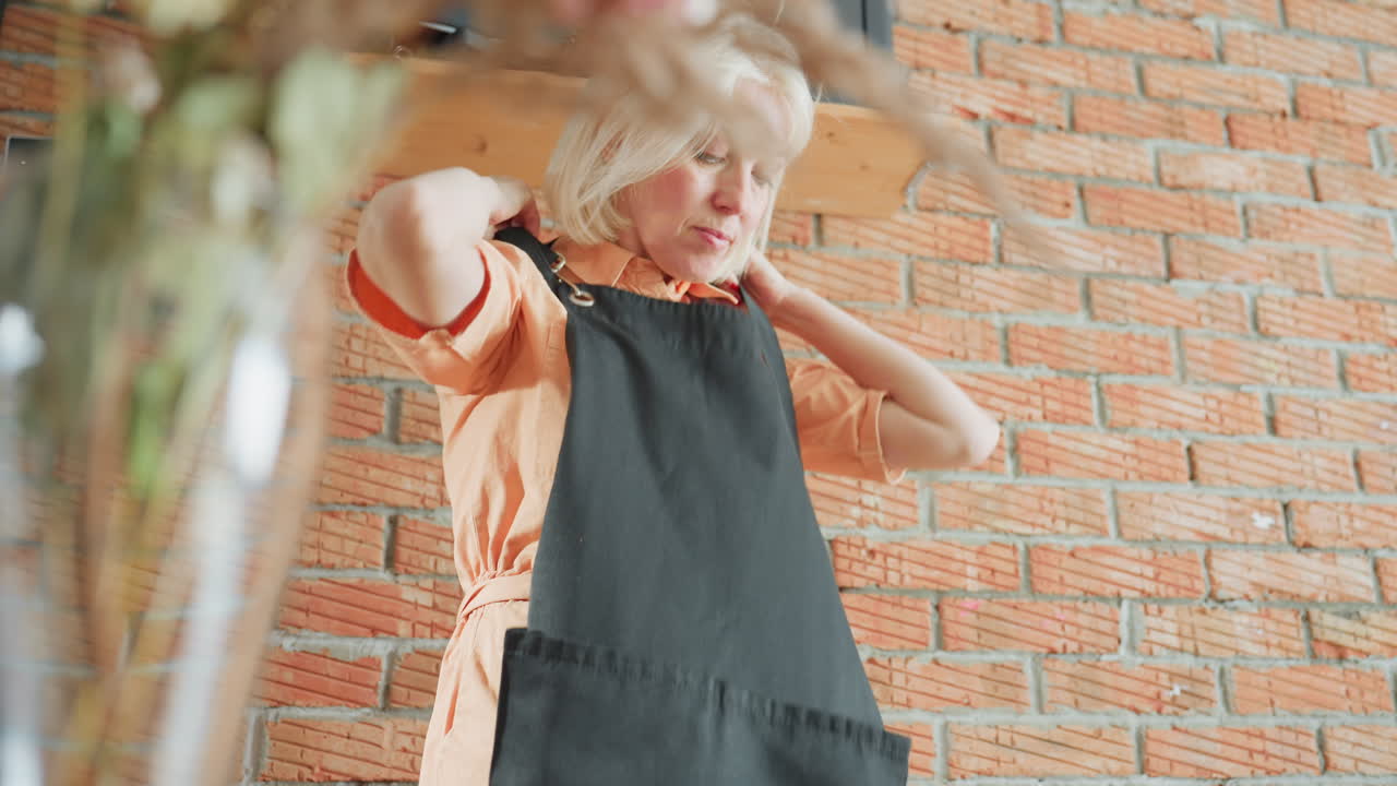 Decorator holding black apron while standing against rustic brick wall, wearing orange shirt, preparing for creative work with focus on readiness, detail, and professional atmosphere in indoor workspace