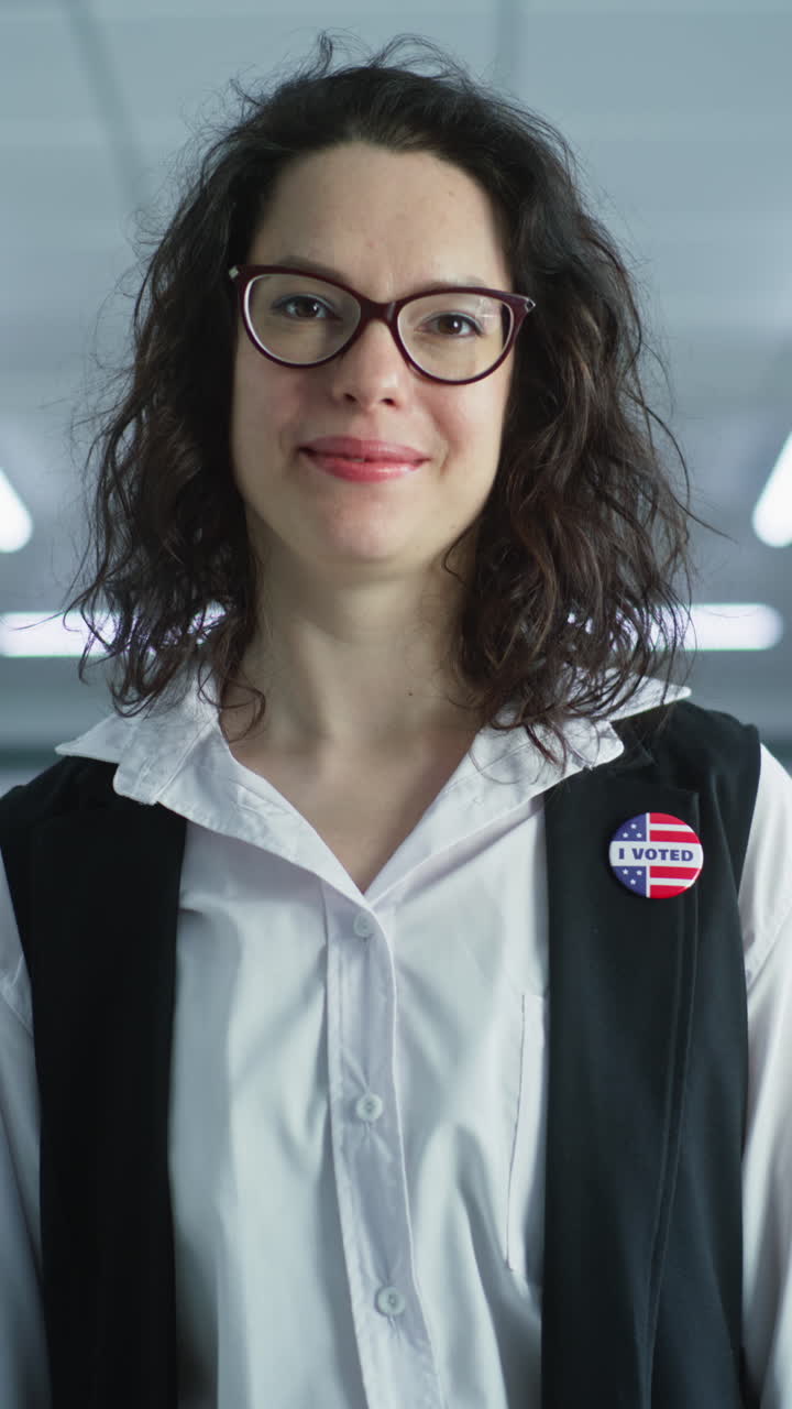 retrato de una mujer caucásica, votante de las elecciones de los estados unidos de américa. una mujer con gafas se para en la estación de votación, posa, sonríe, mira a la cámara. fondo con cabinas de votación. concepto de deber cívico.