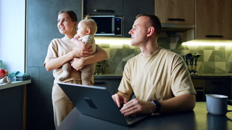 man sitting at the laptop in his kitchen. woman with baby in hands stands behind the man.