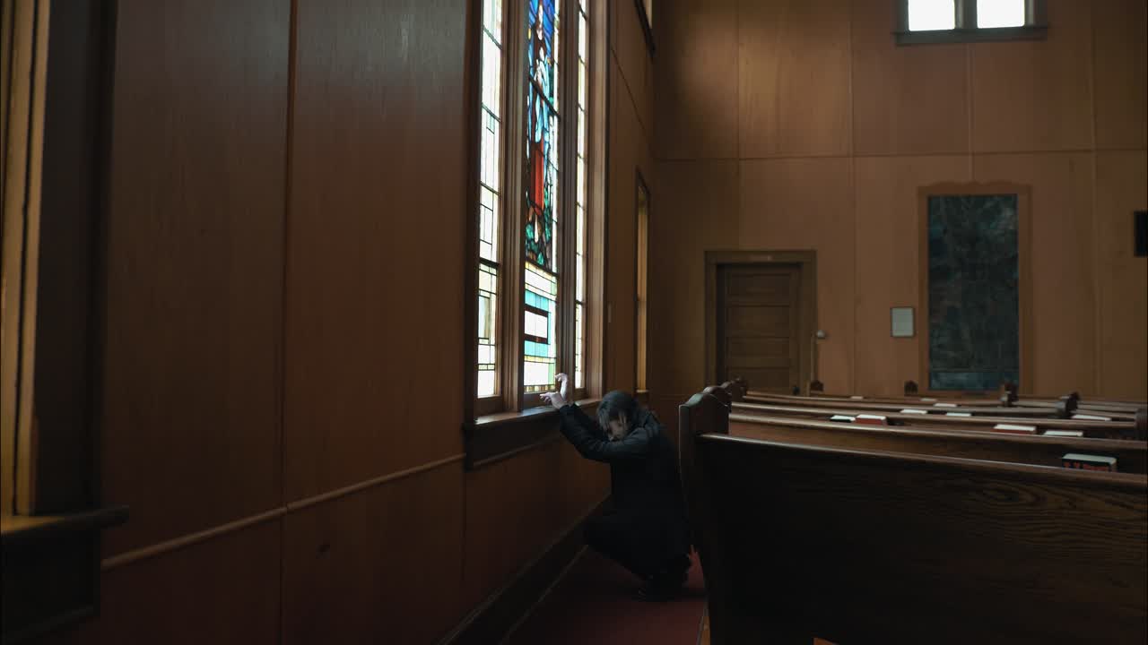 A man in dark clothing gently touches a stained-glass church window, gazing upward in quiet contemplation. The scene conveys spirituality, reverence, and a cinematic mood
