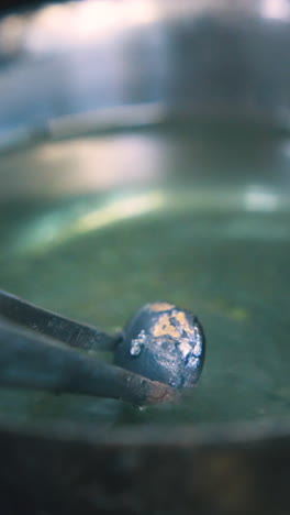 cu: professional jeweler puts metal drop into cup with green liquid using tweezers in dark craft workshop extreme closeup