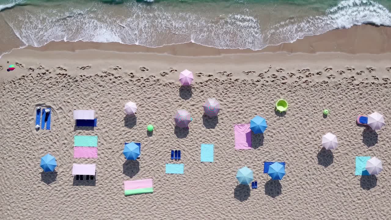 Aerial view of a sandy beach showcasing colorful pastel beach umbrellas and towels, creating a serene and inviting summer scene with the gentle waves lapping at the shore