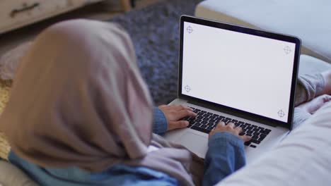 Video-Sobre-El-Hombro-De-Una-Mujer-Birracial-Con-Hijab-En-Casa-Trabajando-En-Una-Computadora-Portátil,-Copiando-Espacio-En-La-Pantalla