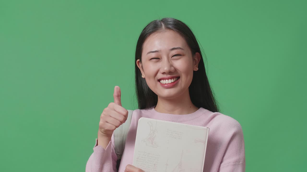 아시아 여성 학생이 책을 들고 녹색 스크린 스튜디오에서 엄지손가락을 보여주고 있습니다.