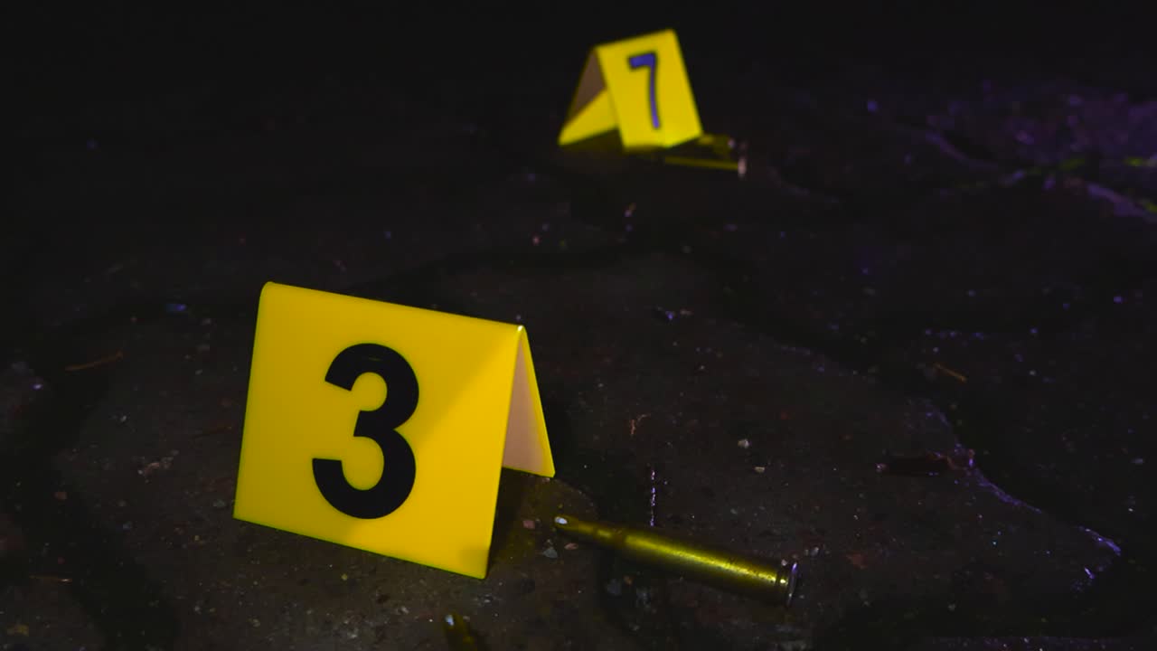 Close up video gliding over yellow colored numbered crime markers next to bullets or gun ammunition on night time asphalt groud in an urban area while red and blue police lights are flashing, lighting