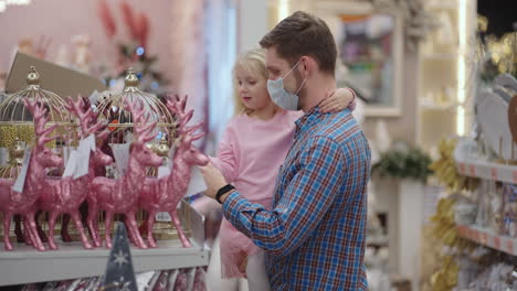 father in medical mask with daughter in shopping mall choose home decorations for christmas