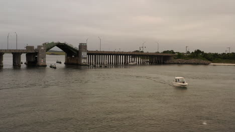 Un-ángulo-Bajo,-Vista-Aérea-De-Un-Pequeño-Bote-Navegando-En-La-Bahía-De-Baldwin,-Más-Allá-De-La-Apertura-De-Un-Puente-Levadizo,-En-Un-Día-Nublado