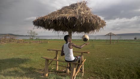 Hinterschuss-Eines-Jungen-Afrikanischen-Mannes,-Der-Sich-An-Einer-Strandhütte-Am-Ufer-Des-Viktoriasees-Entspannt,-Während-Er-Mit-Einem-Fußball-Spielt