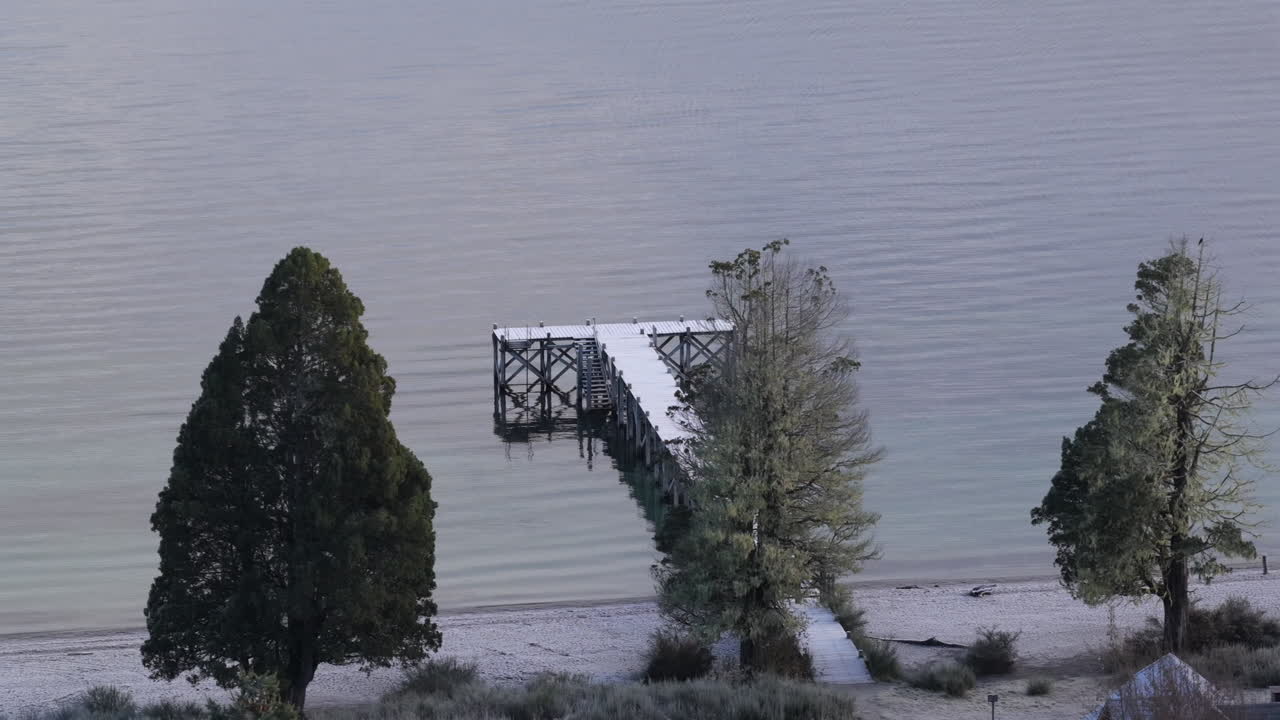 Pontile in legno e riva sabbiosa del Lago Espejo, incorniciati da alberi lacustri vicino a Villa La Angostura, Provincia di Neuquén, Patagonia, Argentina, con la superficie calma del lago e un ambiente naturale tranquillo, rivelati da una lenta ripresa aerea.