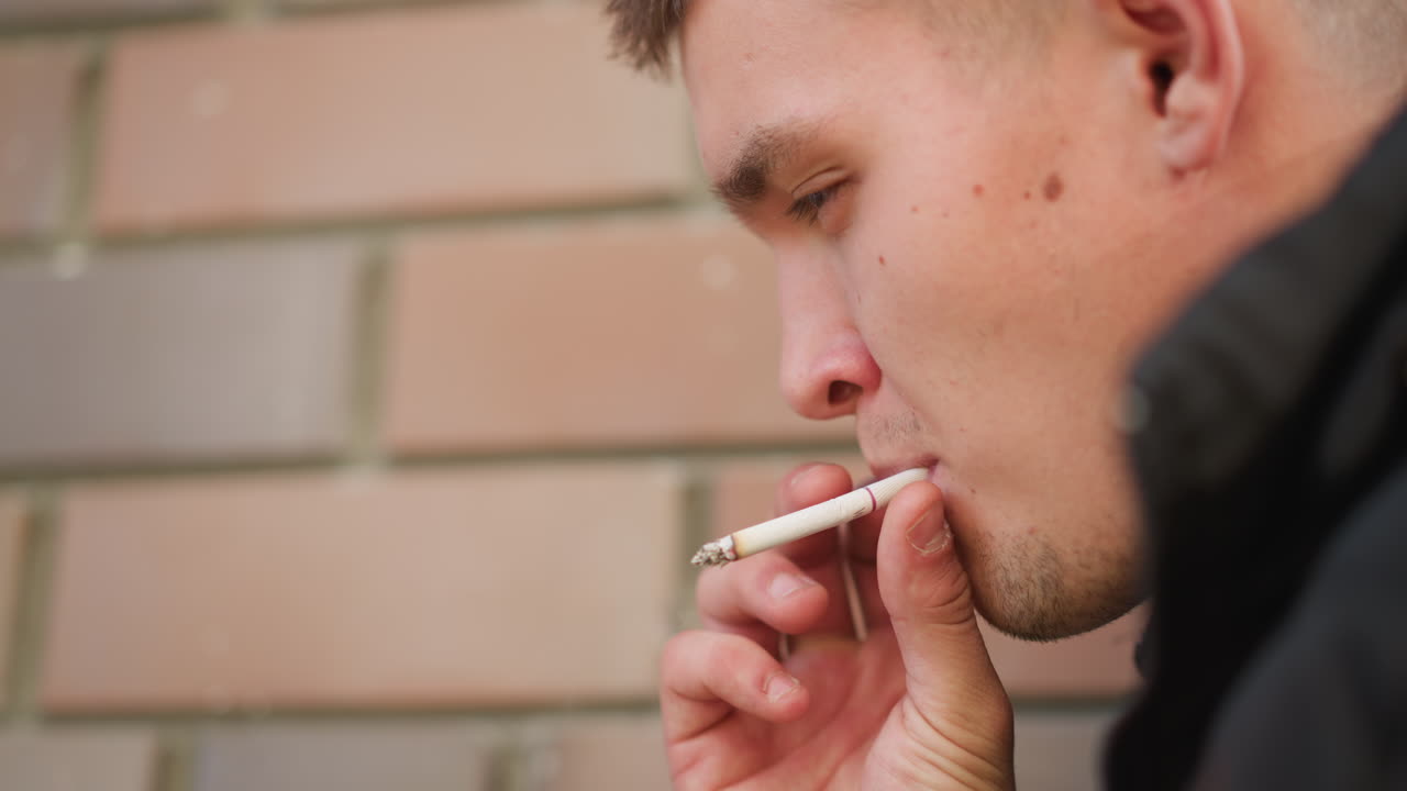 close up of man smoking cigarette while leaning against brick wall, eyes gently closed in calm reflective mood, warm daylight highlighting texture of skin, fingers, and cigarette tip