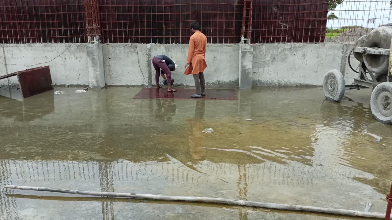 The ground is covered with accumulated rainwater and one person appears to be clearing the water while the other stands nearby.