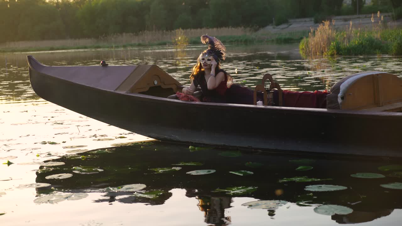 beautiful woman in a dress is floating on a gondola and puting on a carnival mask on the river