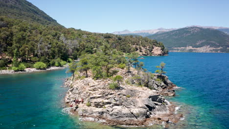 People-jump-into-water-from-islet-by-hilly-coast-in-Argentina,-aerial