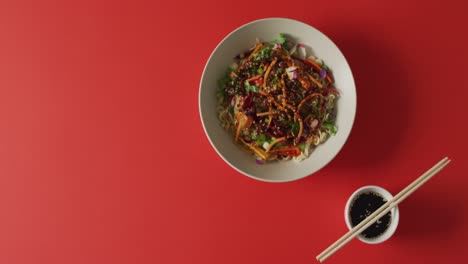 Composition-of-bowl-of-stir-fry-noodels-with-soy-sauce-and-chopsticks-on-red-background