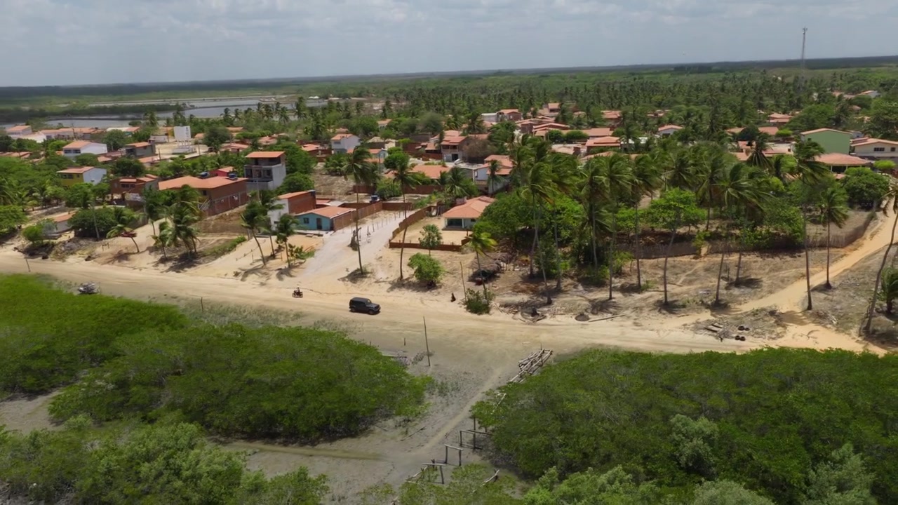 Veículo 4x4 percorre estrada de areia em Guriú, Camocim, Ceará, Brasil, com casas baixas, palmeiras e zonas úmidas estuarinas entre dunas e a costa atlântica, com tomada de drone em acompanhamento panorâmico para plano orbital.