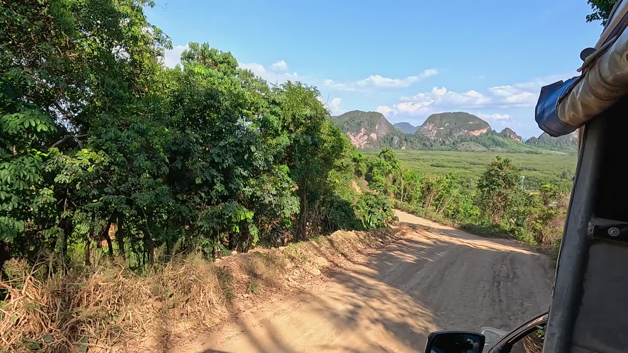 A vehicle navigates a dirt road in Phuket, Thailand, surrounded by lush greenery and distant mountains under clear skies