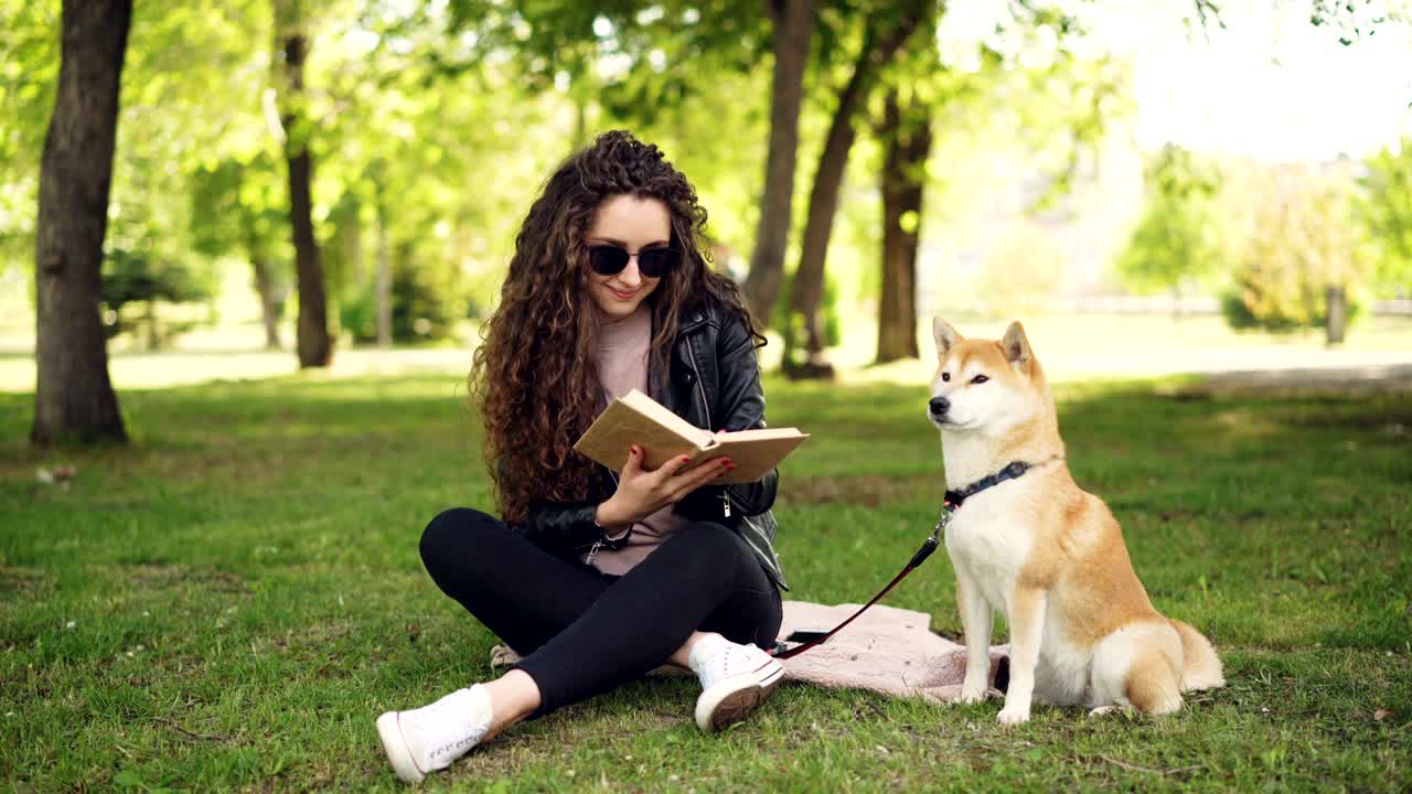 Cheerful female student is reading book in the park sitting on lawn and caressing beautiful dog, pet is yawning and licking its muzzle, girl is laughing.