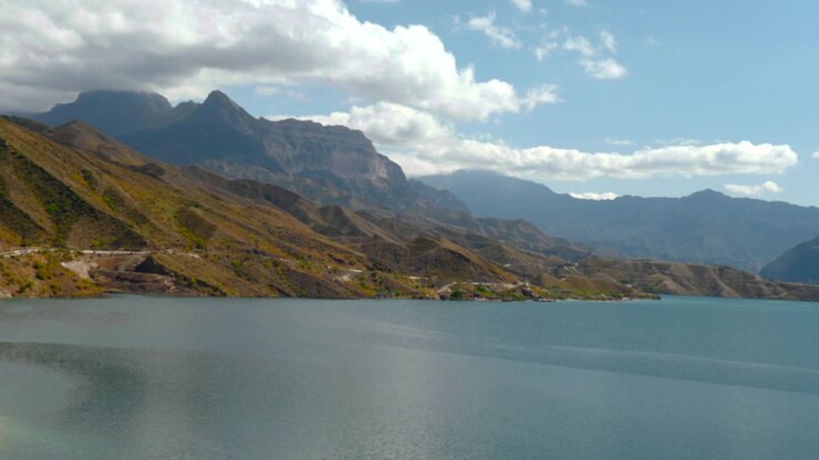 Acqua costiera blu calma riempie il primo piano sotto pendii montuosi scoscesi, creste distanti sovrapposte, una strada costiera tortuosa e un piccolo insediamento sotto la luce del giorno brillante con nuvole soffici, creando un paesaggio sereno