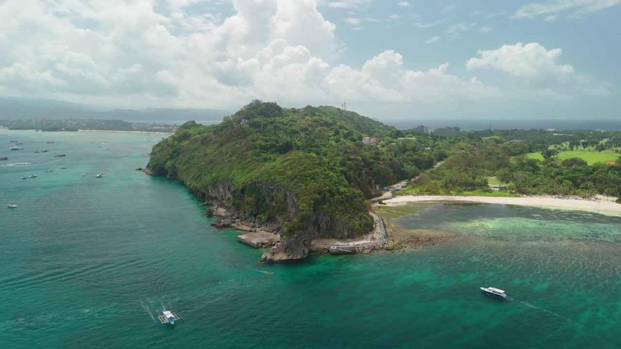 Pengambilan gambar udara mundur dari tanjung batu kapur Cujo's Keyhole di Crystal Cove, Boracay, menampakkan pantai pasir putih, perahu yang ditambatkan, dan garis pantai pulau yang lebih luas, Filipina
