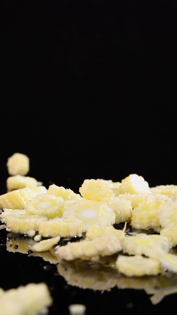 Pezzi di mais baby affettati cadono e si disperdono su una superficie nera lucida, ripresi al rallentatore con illuminazione drammatica e riflessi nitidi.