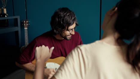 A-brunette-man-in-glasses-in-a-dark-red-t-shirt-plays-the-electric-guitar-for-a-girl-in-a-light-t-shirt-against-a-turquoise-wall