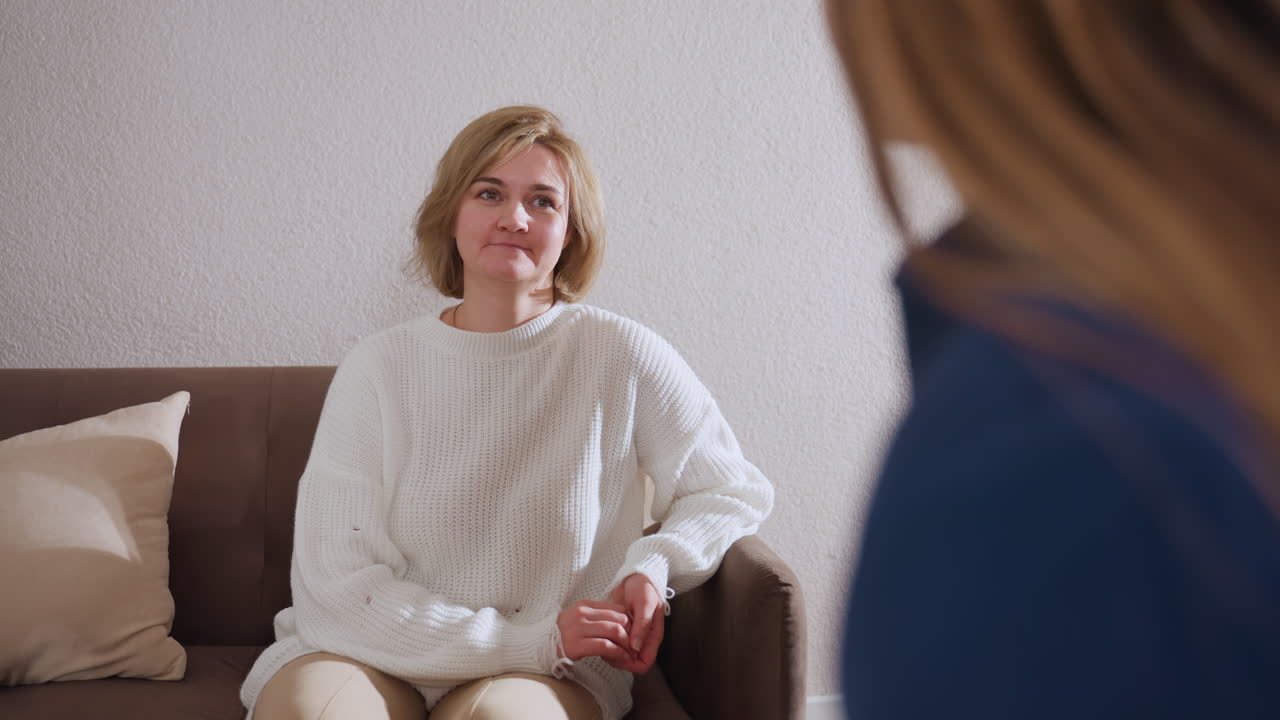 Over shoulder of companion in healing space while cheerful visitor in white sweater smiles gently and listens attentively, reflecting empathy and emotional connection in calm therapeutic setting