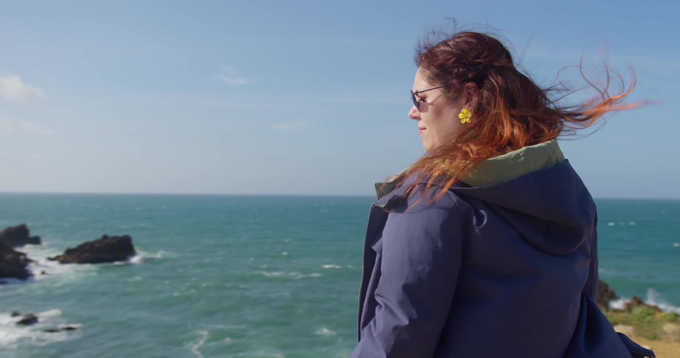 A slow-motion 4K shot of a person in a blue coat watching the stunning Peniche coast, with the ocean crashing against the rocks in a windy, dramatic landscape.