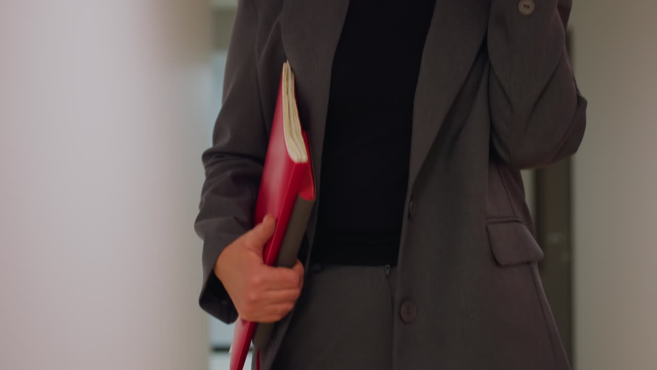 Close-up of businesswoman holding red folder in office corridor, dressed in gray suit, confident professional in modern work environment, career-focused, stylish business attire