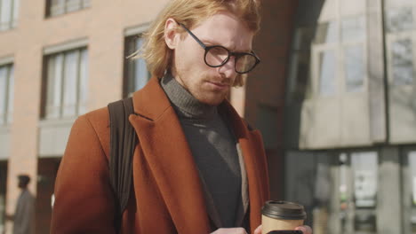 young businessman standing outdoors with coffee and using smartphone