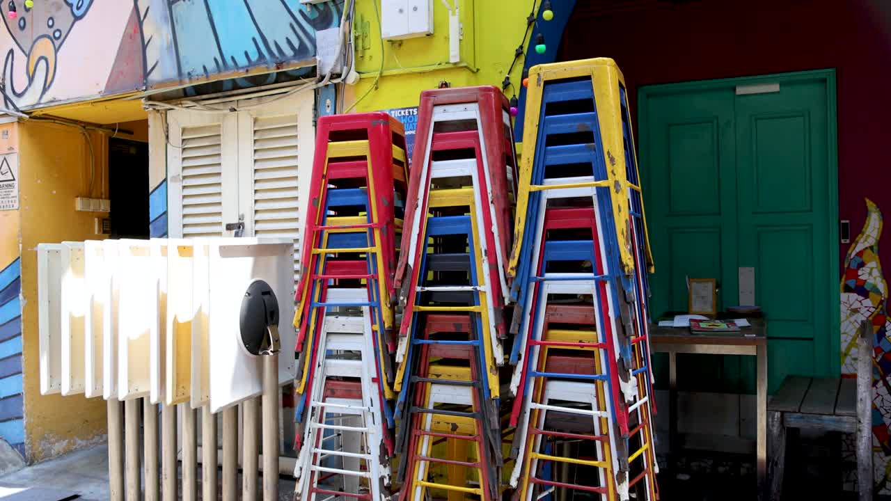 Brightly colored plastic chairs stacked in zigzag patterns outside a vibrant cafe, with sunlight casting strong shadows in a lively urban Singapore alley