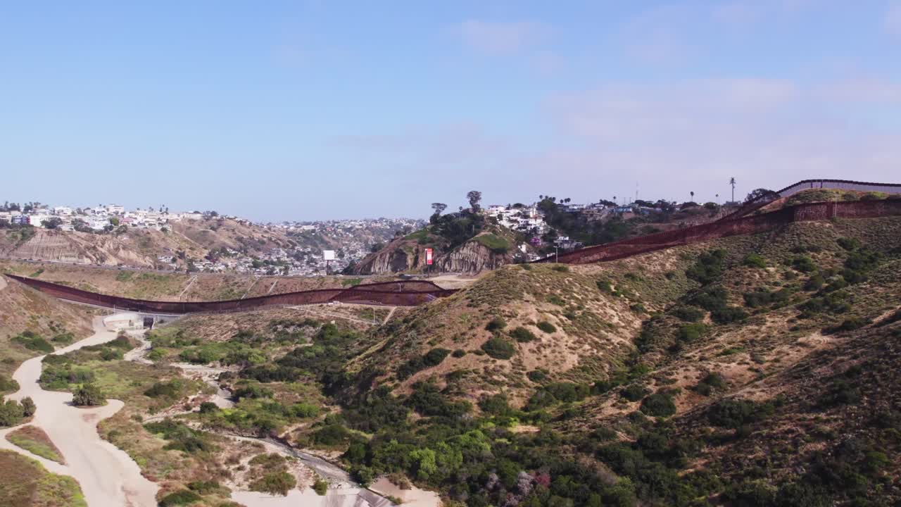 Drone footage captures the winding U.S.-Mexico border wall cutting through rugged hills and desert, with residential areas visible on both sides under a clear blue sky.