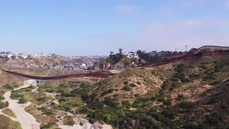 drone footage captures the winding u.s.-mexico border wall cutting through rugged hills and desert, with residential areas visible on both sides under a clear blue sky.