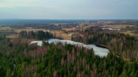 Drones-Aéreos-Vuelan-Sobre-Un-Pequeño-Lago-Congelado-Entre-Pinos-Otoñales-Campo-De-Aldea-Pequeña-Ciudad-En-El-Fondo-Del-Horizonte-Rural,-Paisaje-Otoñal,-Prados