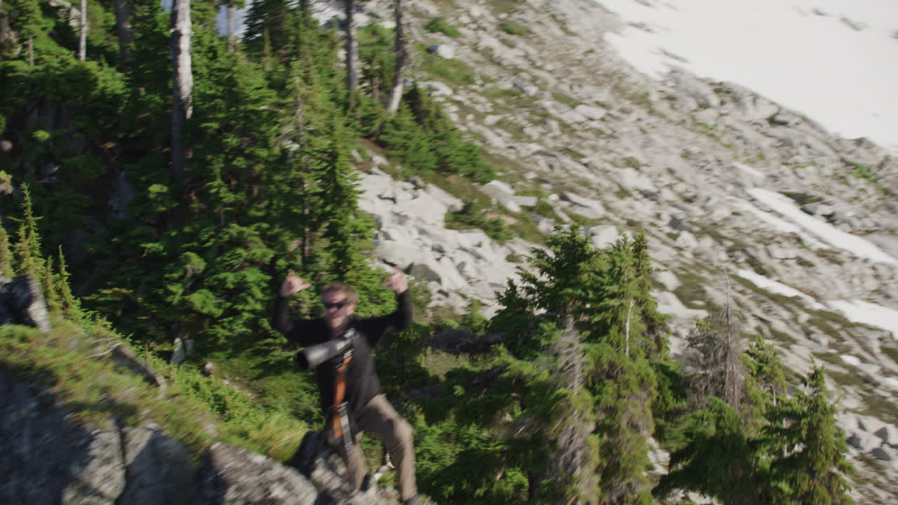 Photographer triumphantly raises both hands and throws peace sign and shaka on mountain ridge, Tim Durkan wide helicopter fly over SLOW MOTION