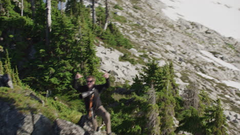 photographer triumphantly raises both hands and throws peace sign and shaka on mountain ridge, tim durkan wide helicopter fly over slow motion