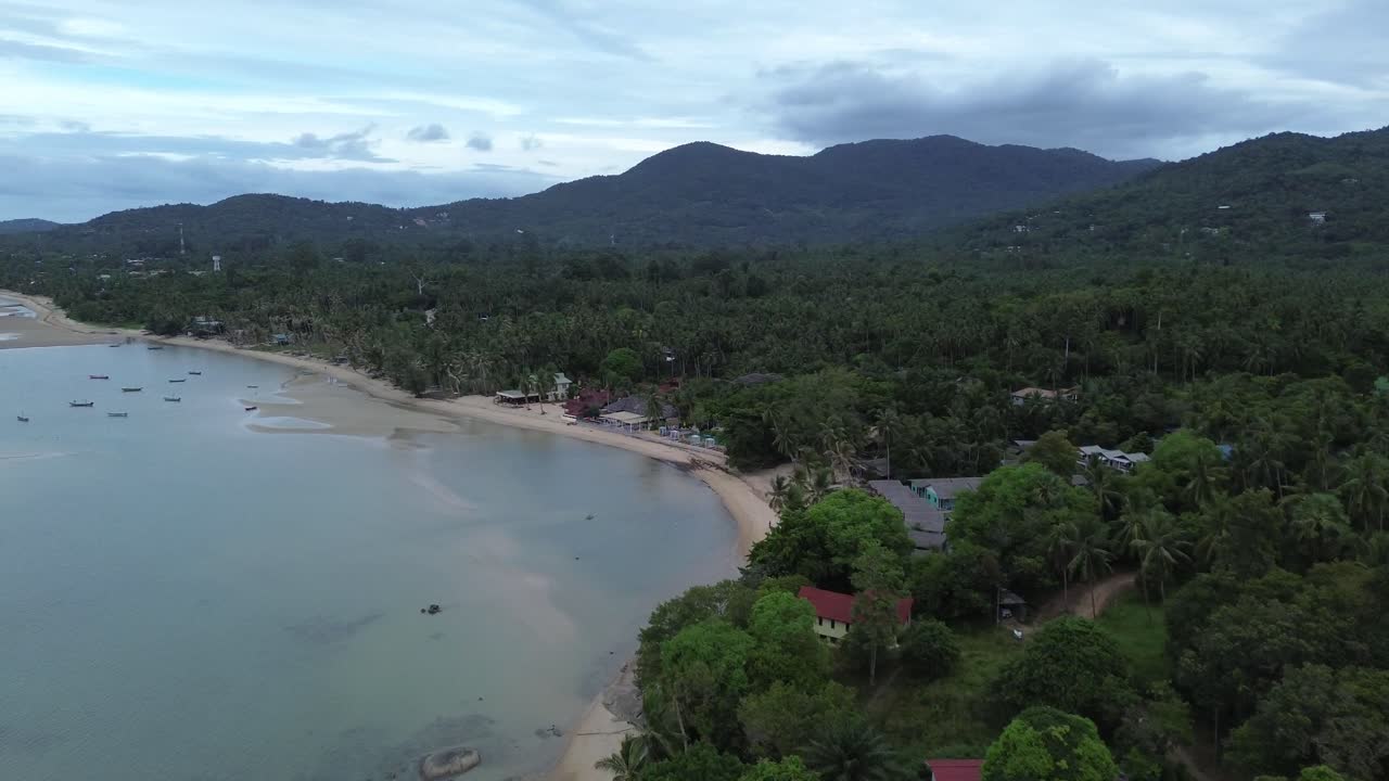 Aerial drone footage of Koh Phangan on a cloudy day, showing tropical coastline, beachfront bungalows and scenic mountains and hills in the background of southern Thailand