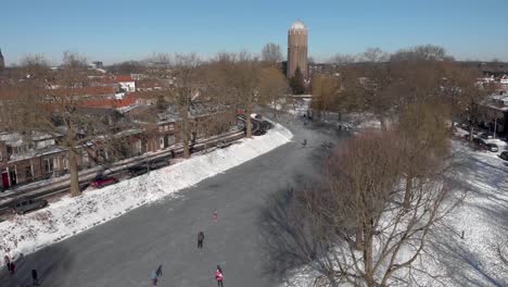 Seitliche-Luftschwenkung,-Die-Eine-Urbane-Winterszene-Mit-Menschen-Zeigt,-Die-Entlang-Eines-Gebogenen,-Zugefrorenen-Kanals-Schlittschuh-Laufen,-Der-Durch-Die-Niederländische-Stadt-Zutphen-Mit-Dem-Ehemaligen-Wasserturm-Im-Hintergrund-Führt