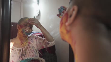 esperanzada paciente de cáncer calva aplica polvo de color en las mejillas en casa frente al espejo en holi, festival indio de colores