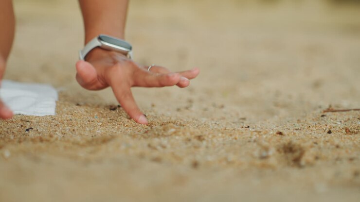 Mani in primo piano che toccano sabbia calda, donna con orologio e braccialetto esplora delicatamente la superficie granulosa della spiaggia con lenti movimenti tattili, texture illuminata dal sole, esercizio di radicamento meditativo, intimo dettaglio macro.