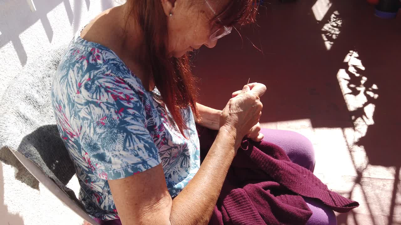 Older Caucasian woman sews her home clothes on a balcony with sunshine, portrait view