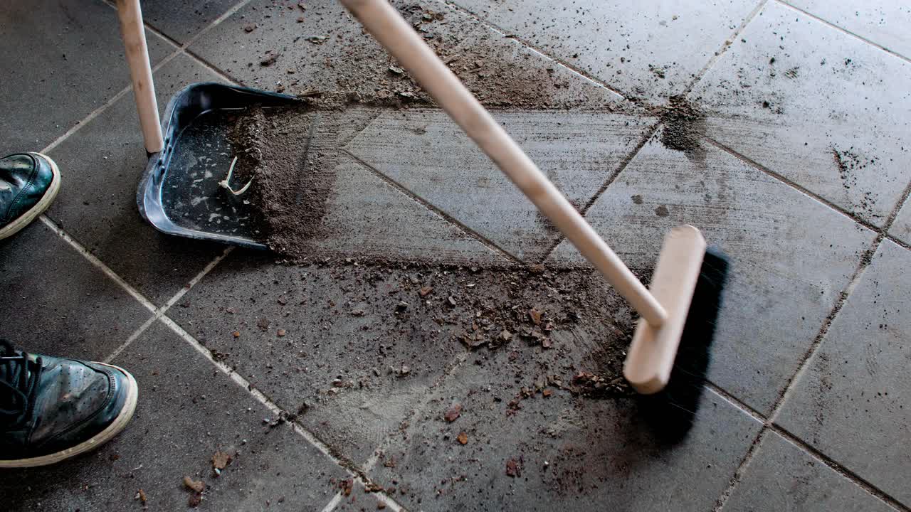 Sweeping a dirty tiled garage floor in a dust pan. Static 4K shot.