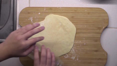 Hands-rolling-thin-dough-with-plastic-bottle-on-wooden-cutting-board,-overhead-shot