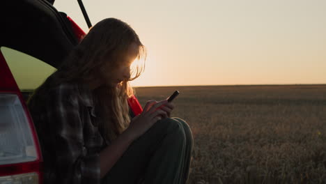 A-teenage-girl-sits-in-the-trunk-of-a-car,-uses-a-smartphone.-Against-the-backdrop-of-a-rural-landscape-where-the-sun-sets