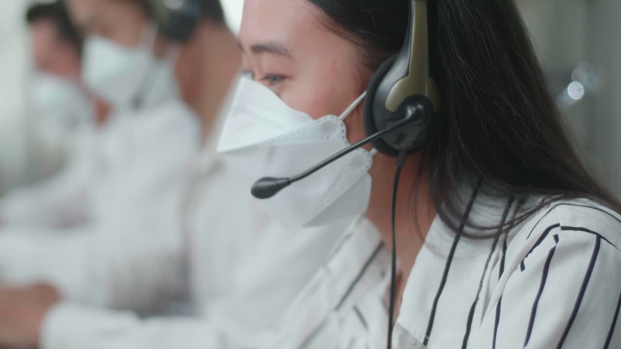 Close Up Of An Asian Woman Call Center Agent Wearing Headset And Mask Typing On Computer While Speaking To Customer On The Call During Working With Colleagues At The Office