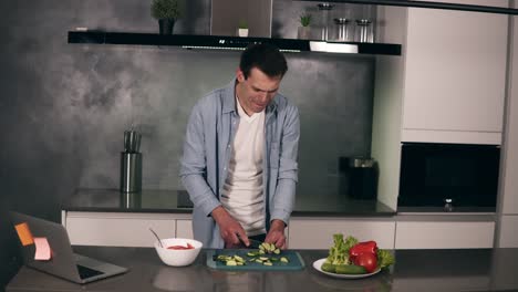 Caucasian-young-man-in-casual-clothes-cooking-vegetarian-breakfast-cutting-vegetables-for-salad-in-kitchen-at-home-and-dancing